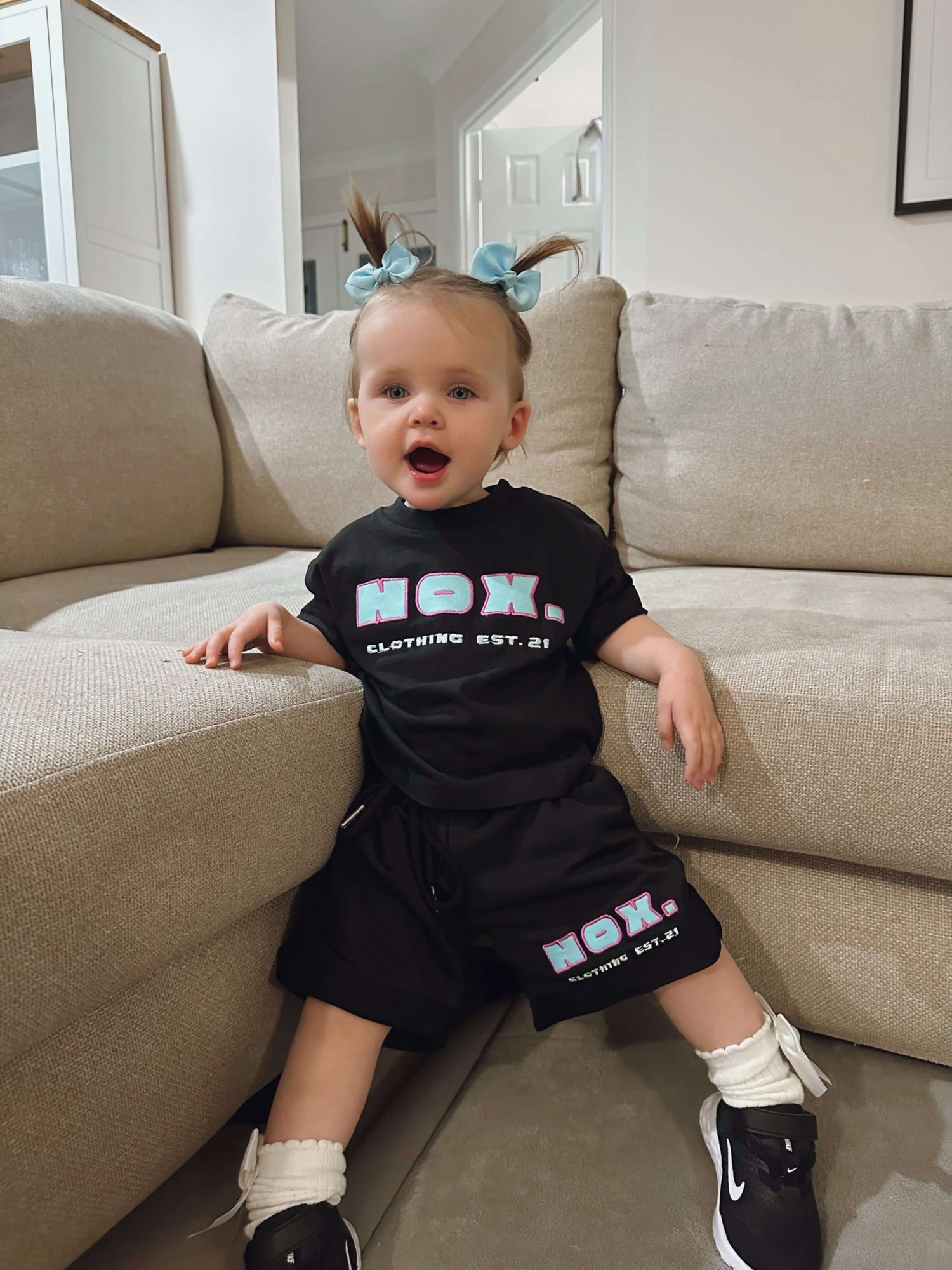 Adorable toddler wearing black NOX Clothing outfit with matching shorts and top, sitting on a beige sofa