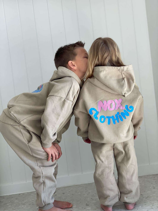 Two children wearing cozy beige fleece logo sets with hoods, showing off "Nox Clothing" branding in colorful letters.